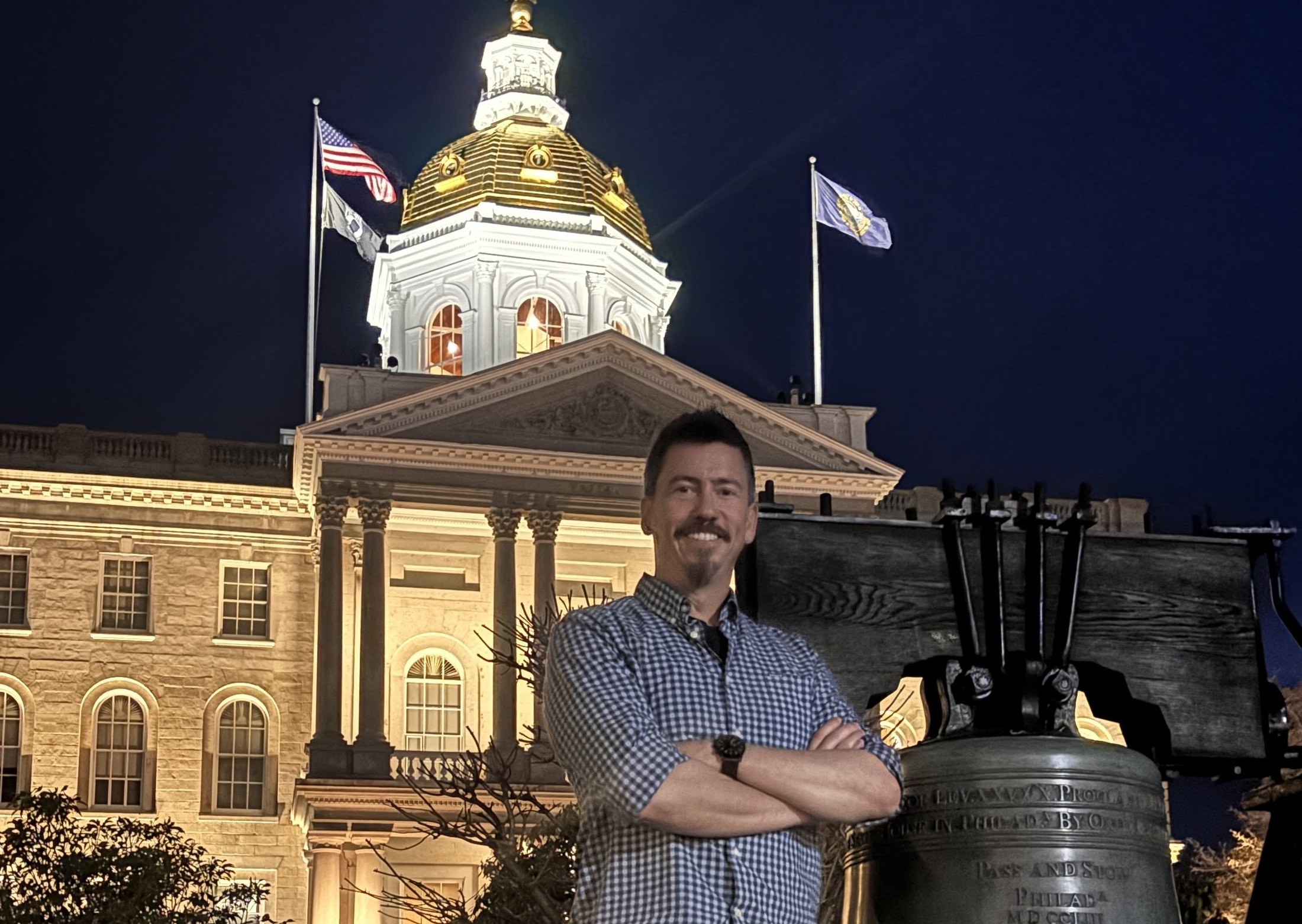 Pedro Serra outside the New Hampshire State House at night, gold dome and replica Liberty Bell behind him